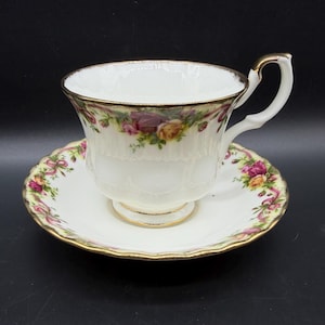 Può includere: Servizio da tè bianco con tazza e piattino con un motivo floreale di rose rosa, gialle e viola. La tazza e il piattino hanno bordi dorati e un bordo smerlato, su sfondo nero.