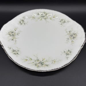 Può includere: Un piatto da torta in ceramica bianca con un bordo ondulato e un motivo floreale con rose bianche e foglie verdi. Il piatto ha un bordo argentato.