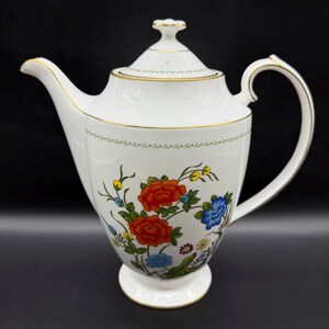 May include: White porcelain teapot with a lid, handle, and spout. The teapot is decorated with a floral design featuring red, blue, and yellow flowers. Gold trim accents the rim, handle, and spout. The teapot sits on a white pedestal base.