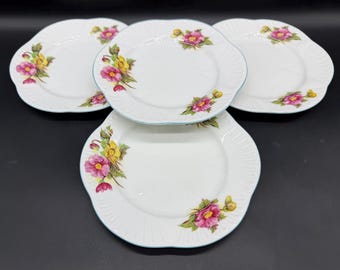 May include: A set of four white ceramic plates with scalloped edges and a ribbed rim design. Each plate features a floral pattern consisting of pink and yellow flowers with green leaves, arranged in two clusters on opposite sides of the plate surface.