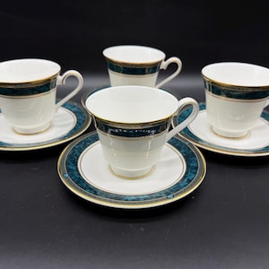 May include: A set of four white teacups and saucers. Each cup and saucer features a teal and gold border. The teacups have a curved handle. The set is displayed on a black surface.