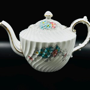 May include: A white teapot with a gold trim and floral design. The teapot has a swirling pattern and features blue and pink flowers.