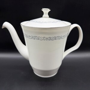 May include: White porcelain teapot with a lid and handle. The teapot has a silver rim and a decorative gray floral pattern around the middle. The spout is curved, and the handle is a simple loop. The lid has a small silver knob.