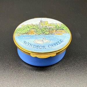Könnte beinhalten: Eine blaue und goldene Schmuckdose mit einem bemalten Deckel mit Windsor Castle, Schwänen und dem Text "Windsor Castle". Die Dose hat einen goldenen Rand und einen blauen Sockel und zeigt eine detaillierte Landschaftsszene.