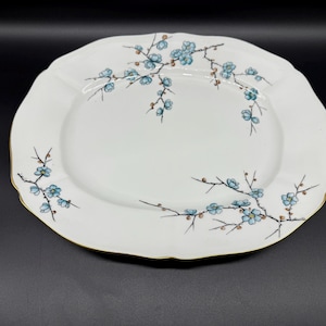 Może przedstawiać: Biała owalna tacka do serwowania z pozłacanym rantem i delikatnym niebieskim wzorem kwiatowym. Kwiaty są stylizowane i przypominają kwiaty wiśni.
