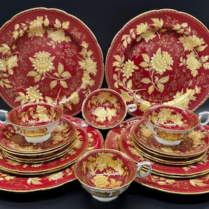 Op de afbeelding: Een set rode en gouden theekopjes en schoteltjes met een bloemenpatroon. De set bevat twee theekopjes, twee schoteltjes, twee theekopjes met schoteltjes en twee grotere borden.