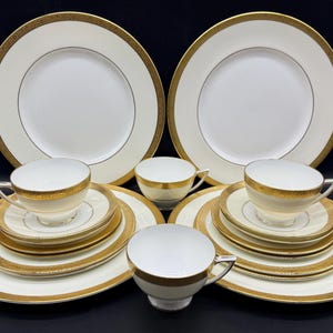 May include: A fine white and gold dinner service. The set comprises large plates, saucers, teacups, and smaller plates. The rims of the plates and cups feature a gold border. The set is arranged on a black surface.