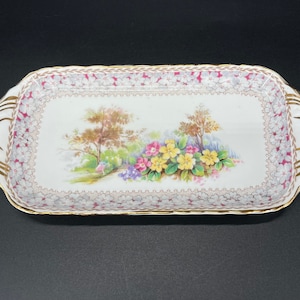 May include: A white porcelain serving tray with gold trim and a pink floral pattern. The tray has two handles and features a painted scene of flowers and trees.
