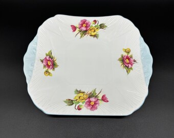 May include: A white square serving plate with a blue rim and floral designs. The flowers are pink, yellow, and green.