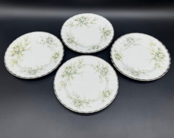 May include: Set of four white ceramic plates with a floral design in shades of green. The plates have a scalloped edge and a black rim.