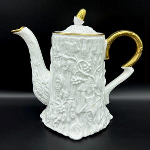 May include: White porcelain teapot with a gold trim and a tree trunk design. The teapot has a lid with a gold acorn finial.