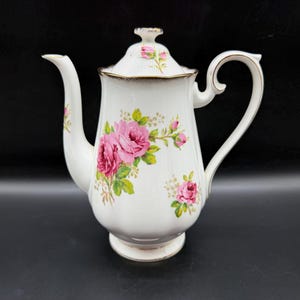 May include: A white porcelain teapot featuring a floral pattern of pink roses with green leaves. The teapot has a scalloped rim, a curved handle, a long spout, and a matching lid with a small finial. Gold trim accents the edges of the lid, rim, and handle.
