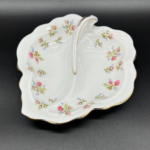 May include: A white porcelain serving dish with a leaf shape and a handle. The dish is divided into two sections and features a floral pattern with pink and blue flowers.