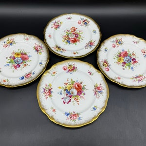 Puede incluir: Cuatro platos de porcelana blanca con ribete dorado y diseños florales. Cada plato presenta un ramo de flores diferente en tonos de rosa, rojo, amarillo y azul.