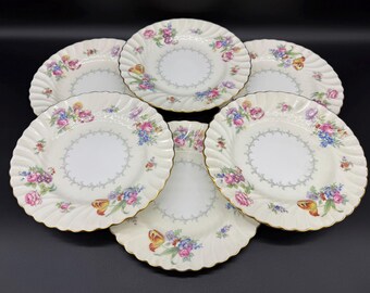 May include: Six white porcelain plates with a floral design featuring pink, blue, and yellow flowers. The plates have a scalloped edge with gold trim and a grey dotted border. The plates are arranged on a black surface.