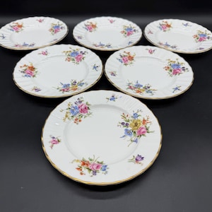 May include: A set of six white porcelain plates with scalloped edges and gold trim. Each plate features a colourful floral pattern with pink, blue, and yellow flowers arranged in clusters around the rim and centre of the white surface.