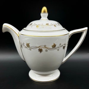 May include: White teapot with a gold lid and gold leaf design. The teapot has a round body and a spout on the left side.
