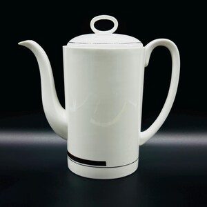 Peut inclure: Une cafetière en céramique blanche avec une bande noire autour du bas. La cafetière a un couvercle avec une petite poignée sur le dessus.