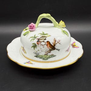 Puede incluir: Un mantequillera de porcelana blanca con una tapa abovedada que presenta un diseño floral con pájaros y mariposas. La tapa tiene un asa de tallo verde con una rosa rosa y una flor amarilla.