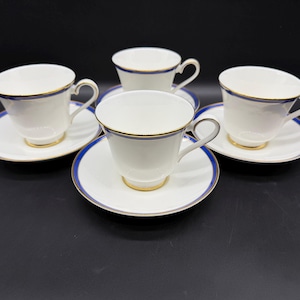 May include: A set of four white porcelain teacups and matching saucers, each featuring a thin navy blue band along the rim and a delicate gold trim. The cups have curved handles and a smooth finish, arranged in a group on a dark surface.