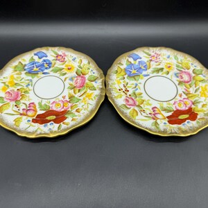 Puede incluir: Dos platillos con decoración floral y borde dorado. Los platillos presentan un centro blanco con una hendidura circular. El diseño floral incluye rosas rosas, campanillas azules y amapolas rojas. Los platillos están sobre una superficie negra.