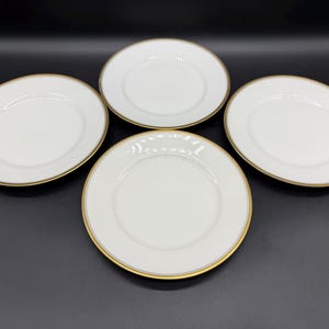 May include: Four white ceramic plates with a gold rim. The plates are arranged on a dark surface. The plates are round and have a simple, elegant design, suitable for serving food.
