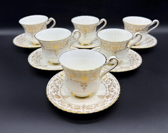 May include: Set of six white teacups and saucers with gold filigree and accents. The teacups have a rounded shape with a handle, and the saucers are round with a matching design. The set is arranged on a dark surface.