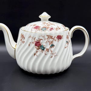 Może przedstawiać: Biały porcelanowy imbryk z spiralnym wzorem i kwiatowym wzorem w kolorze czerwonym, turkusowym i brązowym. Imbryk ma złote wykończenie na uchwycie, wylewce i pokrywce. Klasyczne akcesorium do herbaty.