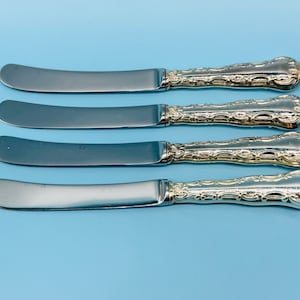 May include: Four silver butter knives with ornate handles. The knives have a polished finish and are arranged in a row on a light blue background.