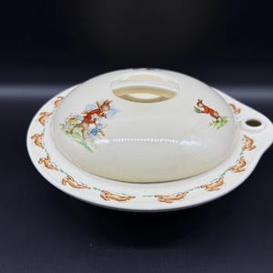 Może przedstawiać: Biała ceramiczna miska do serwowania z pokrywką. Miska ma obwódkę z brązowych królików biegnących wokół krawędzi. Pokrywka ma brązowego królika w czerwonej kurtce i białego królika trzymającego koszyk.