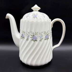 Puede incluir: Tetera de cerámica blanca con un diseño floral en tonos azules y verdes. La tetera tiene un patrón de espiral y una tapa con un pomo.