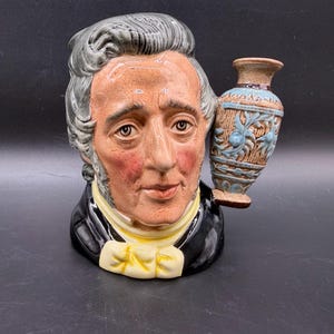 Peut inclure: Figurine en céramique représentant un buste d'homme avec des cheveux gris et un manteau noir. Une petite vase avec des motifs floraux bleus est attachée. Le visage de l'homme est détaillé avec des joues roses et un nœud papillon jaune.