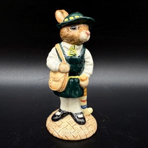 May include: A ceramic figurine of a rabbit dressed in a green hat, vest, and carrying a tan satchel and golf club. The rabbit wears a white shirt and black shoes. The figurine stands on a textured, tan base.