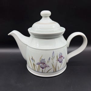 Pode incluir: Um bule de cerâmica branca com tampa de puxador arredondado, alça curva e bico. O corpo do bule é decorado com uma ilustração botânica de flores de íris roxas e folhas verdes que circundam a base do recipiente.