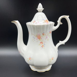 Puede incluir: Tetera de porcelana blanca con un patrón floral y ribete dorado. La tetera tiene una tapa con un pomo y una manija curva.