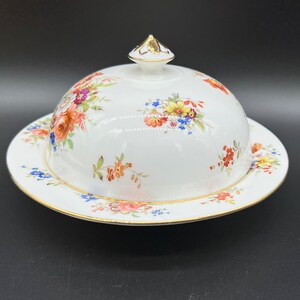 Puede incluir: Un mantequillera de porcelana blanca con un diseño floral. La mantequillera tiene una tapa abovedada con un pomo dorado y una base a juego.