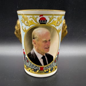 Puede incluir: Un jarrón de cerámica blanca con un retrato de un hombre con traje y corbata negros. El jarrón tiene asas de cabeza de león doradas y está decorado con filigranas doradas y un escudo rojo y dorado. El jarrón mide aproximadamente 13 cm de alto.
