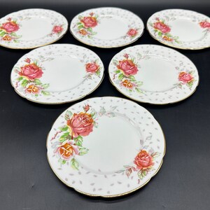 Puede incluir: Seis platos de cerámica blanca con un diseño floral de rosas rosas y un borde dorado. Los platos tienen un fondo blanco con un patrón de lunares gris claro.
