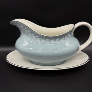 Könnte beinhalten: Eine hellblaue Sauciere mit einem weißen und grauen Blattdesign. Die Sauciere steht auf einer passenden Untertasse.