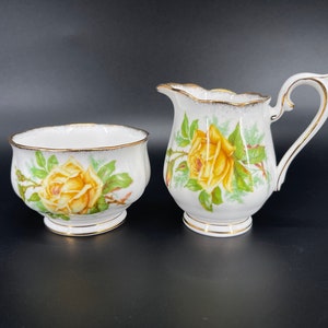 May include: A white porcelain sugar bowl and creamer set with a yellow rose design. The set has a gold trim around the edges.