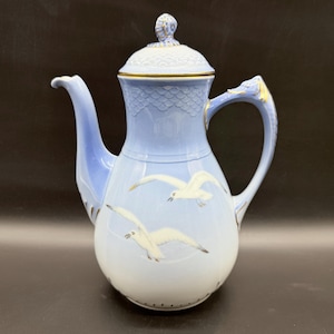 Puede incluir: Una cafetera de porcelana azul claro con tapa y asa. La cafetera presenta un diseño de gaviotas blancas en vuelo. La tapa y el asa tienen detalles dorados. La cafetera mide aproximadamente 25 cm de alto.