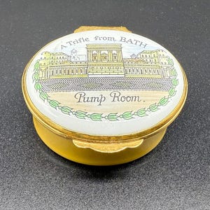 Könnte beinhalten: Eine kleine, goldfarbene Schmuckdose mit weißem Deckel, der eine detaillierte Illustration eines Gebäudes und den Text "A Trifle from BATH" und "Pump Room" zeigt. Die Dose hat einen Klappdeckel.