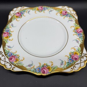 May include: A white, square-shaped decorative plate with a gold rim and floral design. The plate features pink roses, blue and yellow flowers, and green leaves. The rim has a gold filigree design.