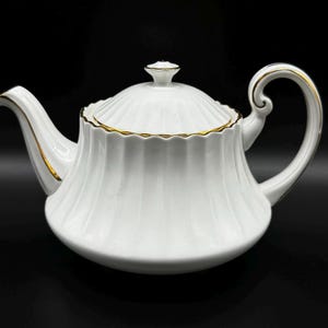 Peut inclure: Théière en porcelaine blanche avec une bordure dorée autour du couvercle et du bec verseur. La théière a un design cannelé et un corps arrondi.