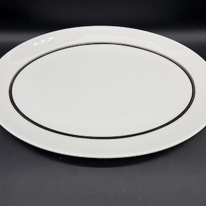 Puede incluir: Una fuente ovalada blanca con un borde negro. El plato tiene un diseño simple y elegante, adecuado para servir aperitivos o platos principales. El plato está hecho de cerámica.