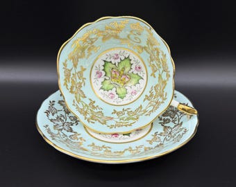 May include: A light blue teacup and saucer set with a gold trim and floral design. The teacup features a green leaf design with a gold crown in the center.