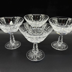 Może przedstawiać: Cztery przezroczyste kieliszki do szampana z kryształu. Każdy kieliszek ma szeroką, płytką misę z teksturowanym wzorem i długą nóżką. Podstawa każdego kieliszka to okrągła stopka z motywem rozety. Kieliszki są ułożone na ciemnej powierzchni.