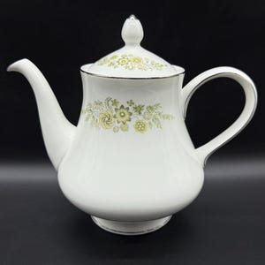 May include: White ceramic teapot with a lid, spout, and handle. The teapot has a floral pattern in green and yellow. The rim and handle are trimmed with silver. The teapot is designed for serving tea.