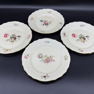 May include: Four white porcelain plates with a scalloped edge and a gold rim. Each plate features a central floral design in pink, blue, yellow, and purple. The plates have a textured basketweave pattern.