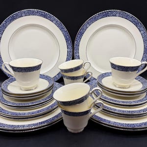 May include: A dinnerware set featuring cream-coloured plates, saucers, and cups with a dark blue floral pattern along the rims. The collection includes multiple stacked plates and matching teacups, all displaying a consistent blue and white design.